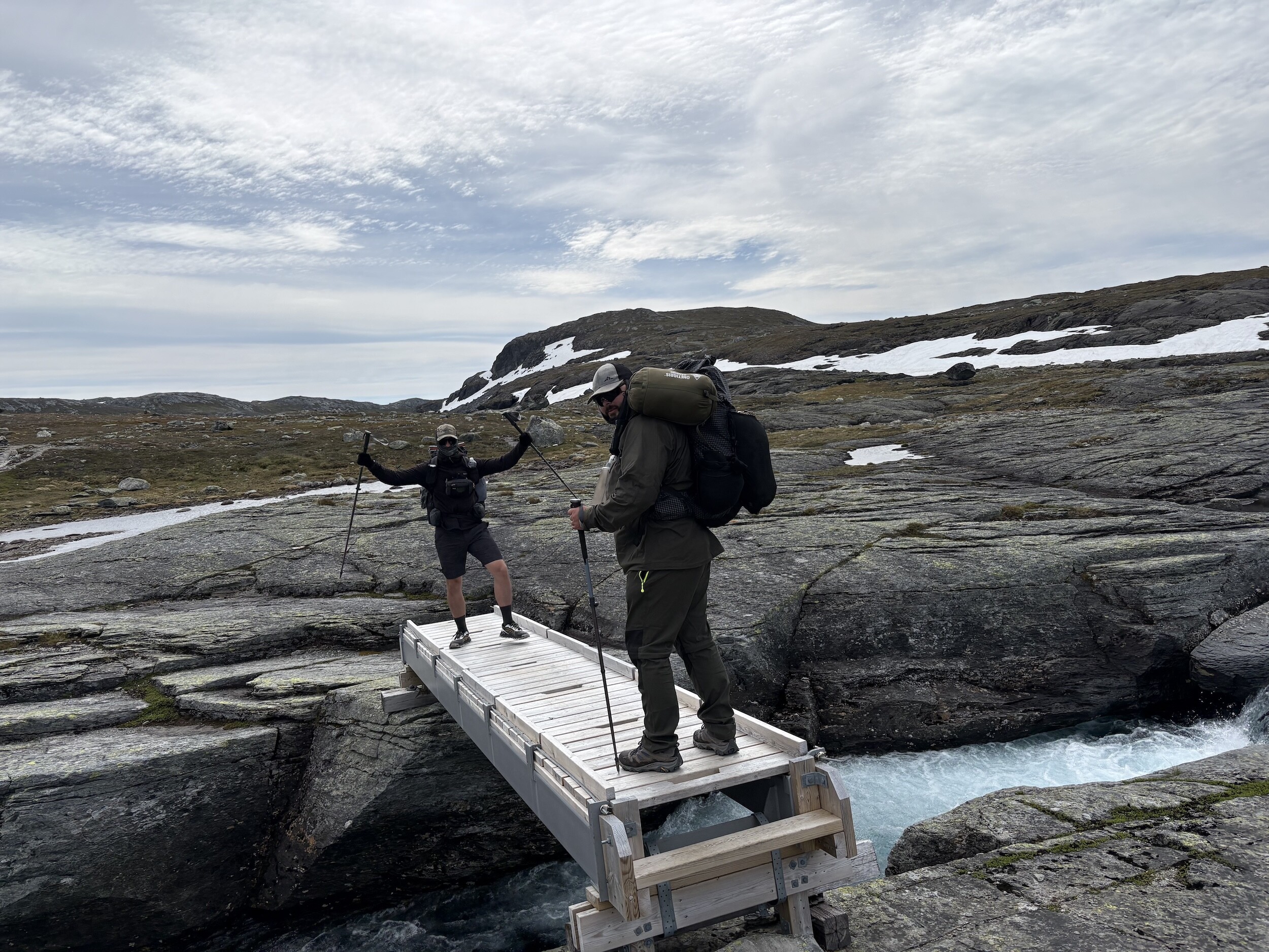Sebastian und Sören mit Trekkingstöcken überqueren eine kleine Brücke über einen Gebirgsbach in der Hardangervidda