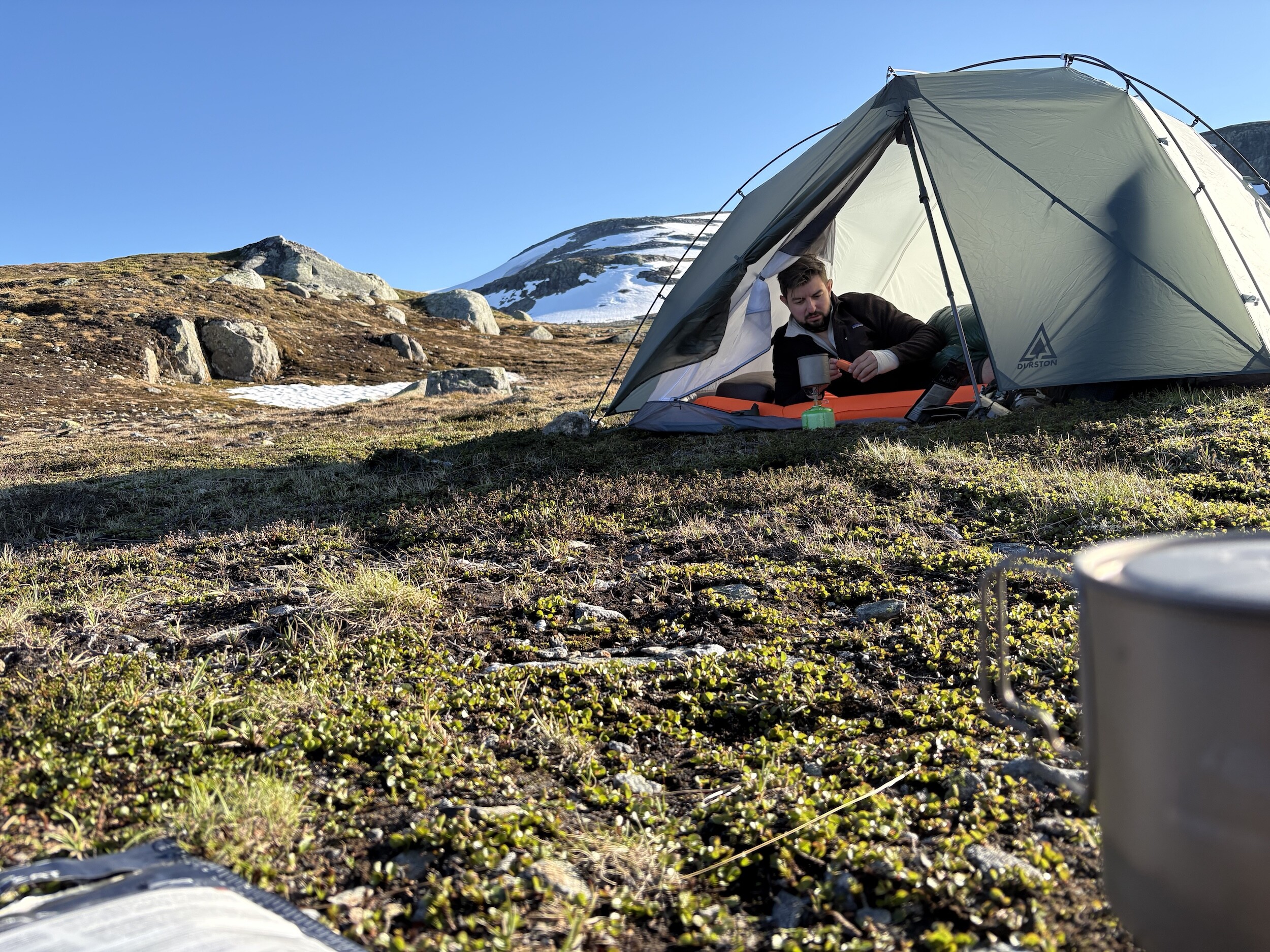 Sebastian bereitet im Zelt auf einer kargen Hochebene in Norwegen Essen zu, mit Schneebergen im Hintergrund